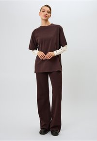 Tummanruskea ylisuuri t-paita, paritettu ribbitettyjen ruskeiden housujen kanssa, joissa on valkoinen pitkähihainen paita mustilla pilkuilla alla; sileä kangas.