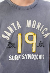 Szürke póló, amelyen a "SANTA MONICA" fehér, a "19" sárga és a "SURF SYNDICATE" fehér színnel látható, hímezett részletekkel és kerek nyakkivágással.