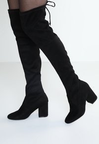 Botas de ante negras hasta la rodilla con punta afilada, tacón cuadrado y detalle de cordones en la parte superior, con una textura suave.