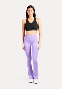 Schwarzer Sport-BH mit rundem Ausschnitt und figurbetontem Design, kombiniert mit hoch taillierten, lila Leggings. Das Model trägt weiße Sportschuhe.