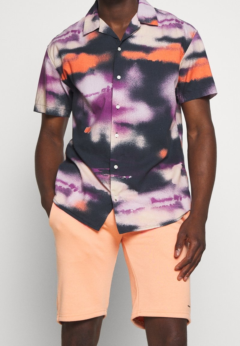 Homme portant une chemise à manches courtes avec un motif tie-dye violet, orange et noir et un short pêche clair.