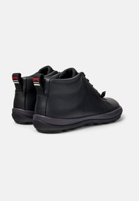 Las botas de cuero negro hasta el tobillo presentan una puntera redonda, suela flexible y una entresuela púrpura contrastante, con tiradores etiquetados con "Camper" en la parte posterior.