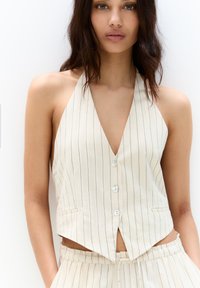 Witte halterneck-top met verticale blauwe strepen, met een diepe V-halslijn, vijf knopen en twee voorkurken. Kort ontwerp.