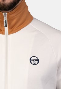 Weißer Zip-Jacke mit einem braunen hohen Kragen, ausgestattet mit einem dunkelblau gestickten Logo auf der linken Brust und einem glatten, strukturierten Stoff.