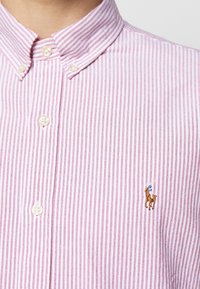 Röd och vitrandig skjorta med krage; har en liten broderad hästlogotyp på bröstet; slät tygtextur.