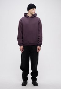 Purple hooded sweatshirt with front pocket, paired with black loose-fitting jeans and black shoes. The model wears a black beanie.