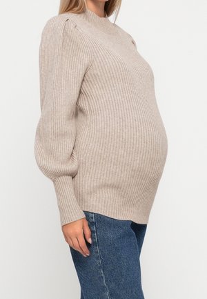 Zwangere persoon die een beige geribbelde trui met lange mouwen en een blauwe spijkerbroek draagt, staand tegen een effen witte achtergrond.