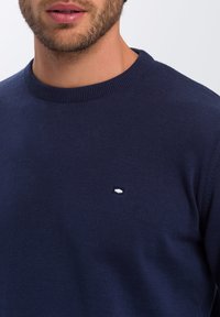 Granatowy sweter dzianinowy z okrągłym dekoltem. Posiada małe białe logo na piersi. Gładka faktura, dopasowany krój.