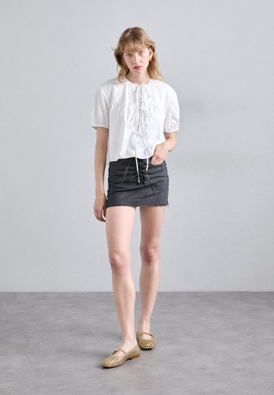 Blusa blanca de manga corta con lazo en el frente, combinada con una falda mini de mezclilla negra con detalles de cordones. Se lleva con zapatos planos estampados.
