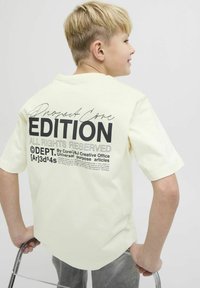 Camiseta amarilla de algodón con un gran texto impreso en negro en la parte posterior. Mangas cortas y un corte relajado. Pantalones grises en primer plano.