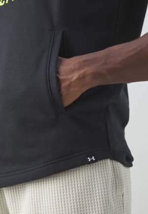 Ciemna koszula z ręką w przedniej kieszeni, białe teksturowane spodnie oraz mała czarna metka z logo Under Armour na dole koszuli.