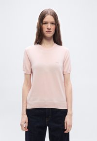 Γυναίκα με μακριά καστανά μαλλιά που φοράει ανοιχτό ροζ πουλόβερ με κοντά μανίκια και σκούρο μπλε τζιν παντελόνι, στέκεται μπροστά σε απλό λευκό φόντο.