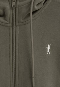 Felpa con zip color khaki realizzata in tessuto morbido, con un piccolo logo ricamato di un golfista bianco sul lato sinistro del petto. Cappuccio regolabile con cordini.