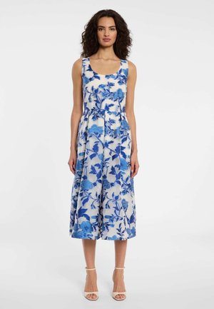 Femme aux cheveux bouclés et foncés portant une robe midi blanche sans manches à imprimé floral bleu et des talons blancs ouverts, debout devant un fond uni.