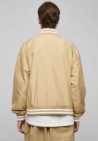 Giacca bomber beige realizzata in tessuto morbido, con polsini e orlo a coste a righe. Vestibilità comoda con un design semplice e senza marchi visibili.