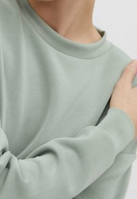 Sudadera verde claro con una textura suave, con cuello redondo y puños ajustados. La tela parece suave y cómoda.
