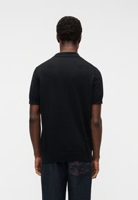 Άνδρ ας που στέκεται γυρισμένος προς τα πίσω, φοράει μαύρο κοντομάνικο πουκάμισο και σκούρο τζιν με περίγραμμα κόκκινου πουλιού στην πίσω τσέπη.