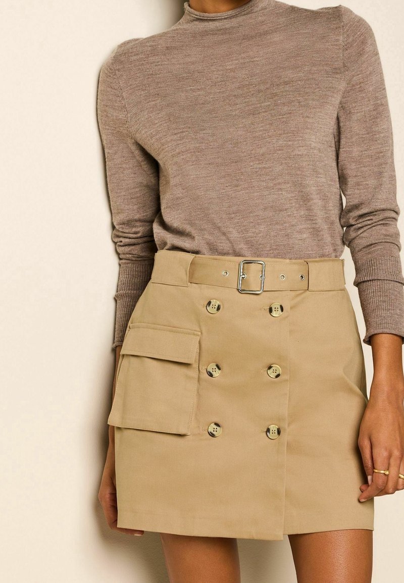 Jupe en coton beige avec un design à double boutonnage et une poche latérale, associée à un haut en maille taupe à manches longues.