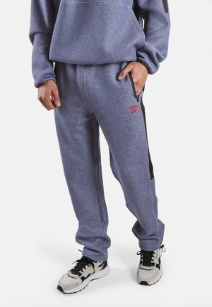 Persoon draagt lichtpaarse Reebok joggingbroek met zwarte zijstreep, hand in zak, gecombineerd met beige en zwarte sportschoenen.