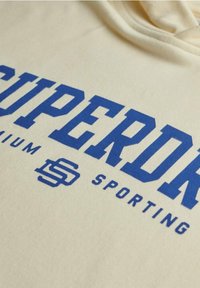 Sweat à capuche de couleur crème avec l'inscription "SUPERDRY" en bleu vif dans un design sportif. Texture douce avec des détails de logo distinctifs.