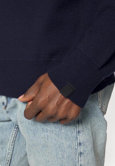 Pull en tricot bleu marine avec des poignets côtelés, une texture lisse et une petite étiquette de marque noire sur le poignet gauche. Porté avec un jean en denim bleu clair.