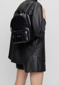 Czarna skórzana plecak o zaokrąglonym kształcie, z zamykaną na zamek przednią kieszenią i wytłoczonym logo. Teksturowana powierzchnia doskonale komponuje się z wzorzystym czarnym strojem.