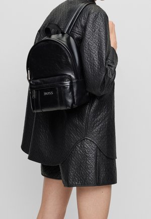 Mustaka nahkareppu pyöreällä muotoilulla, vetoketjullinen etutasku ja kohokuvioitu logo. Teksturoitu pinta täydentää kuviollista mustaa asua.