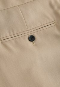 Beige Stoff mit glatter Textur, versehen mit einer Tasche mit einer horizontalen Naht und einem schwarzen Knopf mit vier Löchern für den Verschluss.