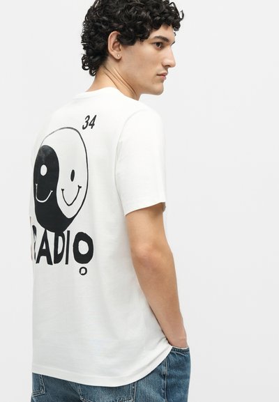Νεαρός άνδρας με κατσαρά μαλλιά φορεί λευκό T-shirt με ασπρόμαυρο γραφικό γιουίν-γιανγκ με χαμογελαστό πρόσωπο και τον αριθμό 34 στην πλάτη, σε συνδυασμό με μπλε τζιν.