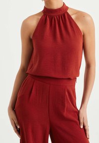 Rostfärgad ärmlös topp med hög hals, texturerat tyg och samlade detaljer. Matchas med högmidjade byxor i samma färg.