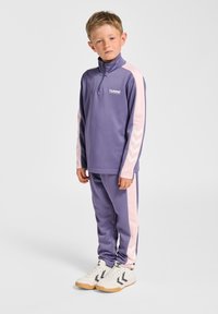 Lilla atletisk træningsdragt med halvlille lynlås i toppen og lyserøde detaljer; har chevronmønster på ærmerne. Bæres med hvide sneakers med beige sål.