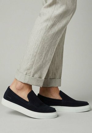 Mann trägt beige hochgekrempelte Hosen und schwarze Slip-on-Sneaker mit weißen Sohlen, steht auf einem hellgrauen Boden.