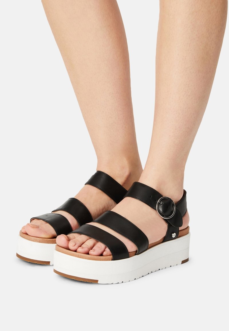 Pieds portant des sandales à plateforme en cuir noir avec trois brides et une boucle ronde argentée sur une semelle blanche sur fond uni.