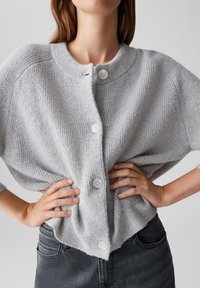 Femme portant un cardigan en tricot côtelé gris clair avec de grands boutons blancs, les mains sur les hanches, associé à un jean gris foncé.