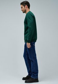 Felpa verde con collo a giro, abbinata a jeans di denim blu. Scarpe scure completano il look. Il modello è posato di lato contro uno sfondo grigio.