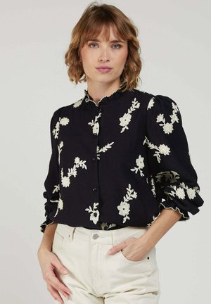 Femme aux cheveux courts châtain clair portant un chemisier noir avec une broderie florale blanche et un pantalon blanc taille haute, main dans la poche.