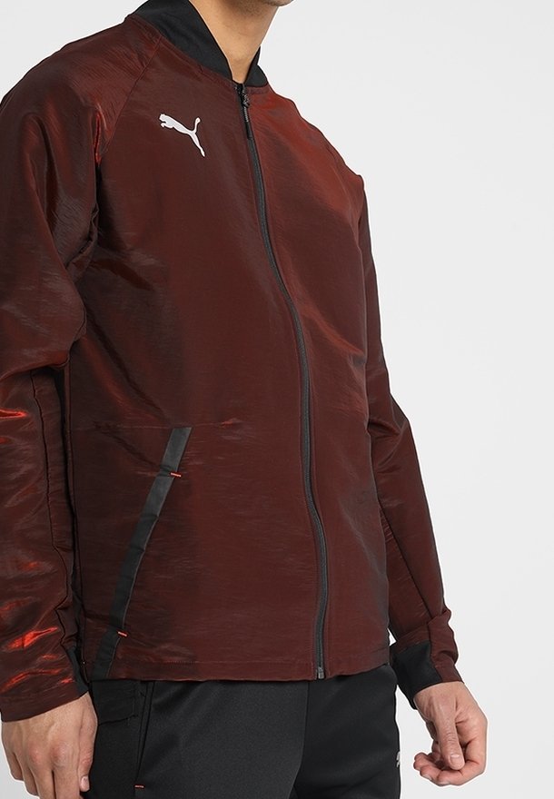 Mężczyzna w bordowej kurtce Puma na zamek z czarnym kołnierzem i kieszeniami wykończonymi czarną lamówką, w połączeniu z czarnymi spodniami.