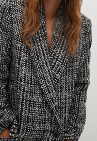 Veste à motif pied-de-poule noir et blanc avec un col cranté, un design croisé à double boutonnage et un seul bouton noir à l'avant.