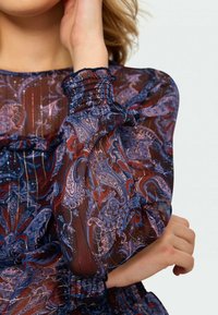 Žena v dlouhém rukávu průhledné blůze s modro-červeným paisley vzorem a nařasenými manžetami, ruka se dotýká její tváře a paže má zkřížené.