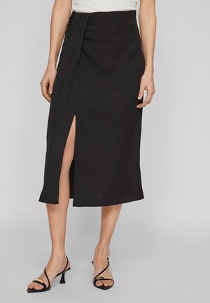 Jupe midi noire en tissu texturé, avec une fente sur le côté et des détails drapés à la taille. Associée à des talons noirs à brides.