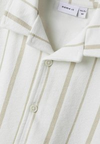 Camisa blanca de cuello con rayas verticales beige, hecha de tejido texturizado. Presenta un cierre de botones y una etiqueta en el interior del cuello.