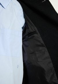 Svart kappa uppvisad öppen, som visar en ljusblå skjorta innanför. Kappan har en texturerad utsida och en slät svart foder.