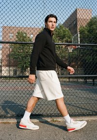 Young man in black jacket and white shorts walking beside a chain-link fence around an outdoor basketball court on a sunny day.