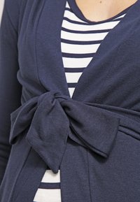 Mörkblå cardigan med en rosettdetalj vid midjan, lager på lager över en vit och mörkblå randig topp. Mjuk, stickad textur, åtsittande design.