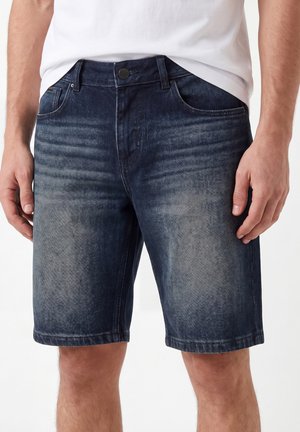 Hombre que lleva pantalones cortos vaqueros azul oscuro con bolsillos delanteros y una camiseta blanca, mostrado desde la cintura hasta las rodillas.