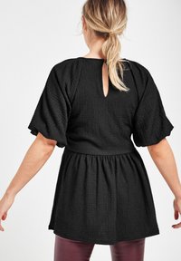Tunique noire texturée avec des manches courtes bouffantes, présentant un dos en forme de trou de serrure et une taille froncée. Associée à des leggings bordeaux brillants.