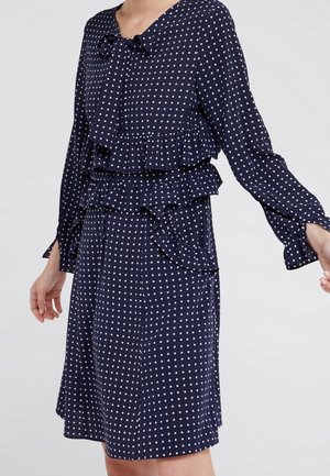 Granatowa sukienka w białe groszki, z długimi rękawami, falbanami przy talii i na biuście oraz kokardą przy dekolcie.