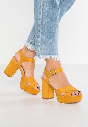 Füße in senfgelben Plateau-Pumps mit Knöchelriemen und Schnalle, kombiniert mit hellblauen Jeans mit ausgefransten Säumen.