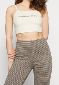 Krémový crop top s logem Calvin Klein Jeans, s tenkými ramínky a měkkou texturou, doplněný o kalhoty s vysokým pasem v černobílém kostkovaném vzoru.