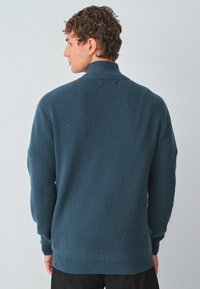 Maglione lavorato a maglia in teal scuro con colletto alto, maniche lunghe, texture a coste e polsini aderenti; design semplice senza decorazioni visibili.